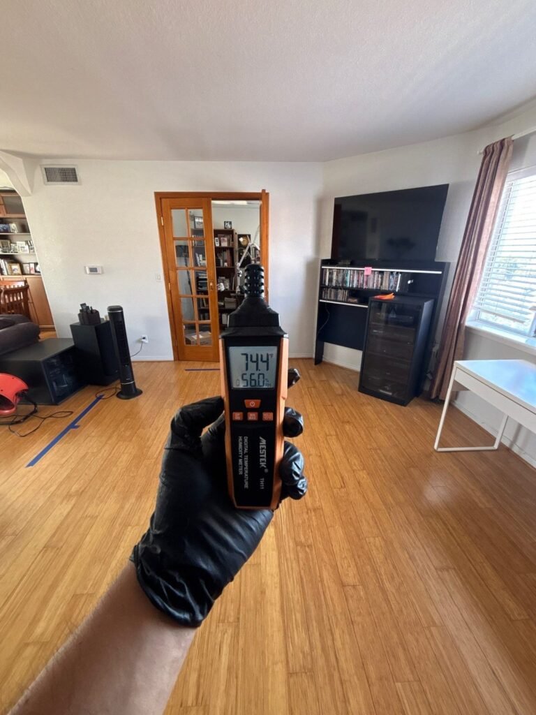 A gloved hand holding a meter reading 74.4 in a furnished living room in the East Bay